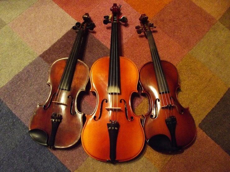 Three stringed instruments: two violins and a cello, resting on a patterned carpet.