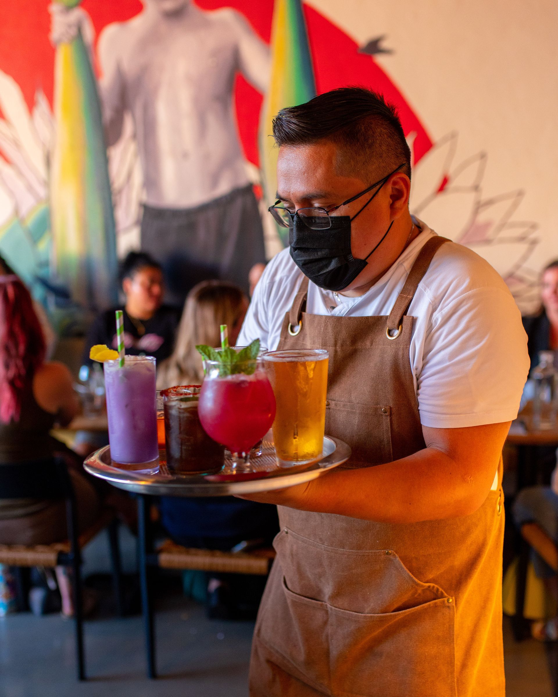 A waiter wearing a mask is carrying a tray of drinks in a restaurant.