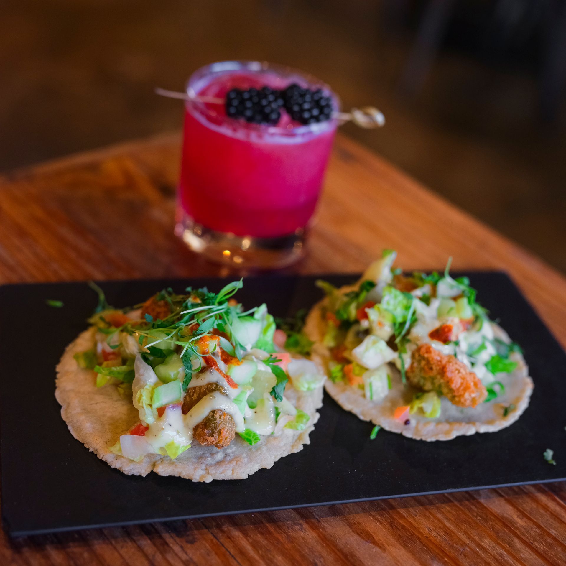 Two tacos on a black plate next to a drink on a wooden table.