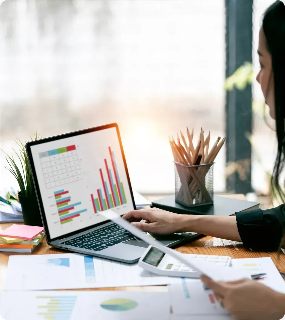 Woman reviewing financial charts on a laptop at a desk with paperwork and calculator.