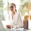 Uma mulher usando óculos está sorrindo enquanto está sentada em uma mesa com um laptop