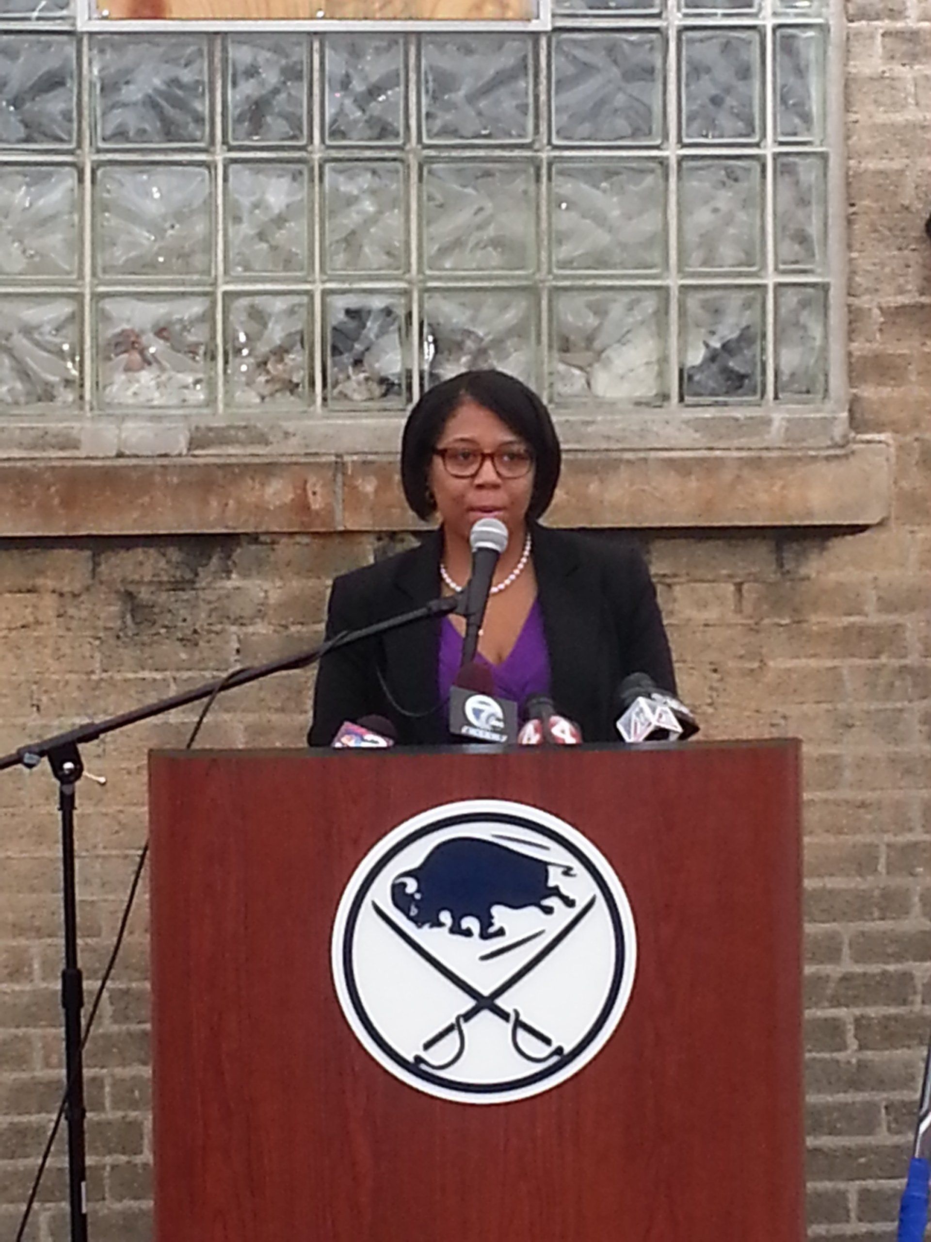 Woman speaking at a podium with Buffalo Sabres logo. Wearing glasses, black blazer, purple top, brick background.