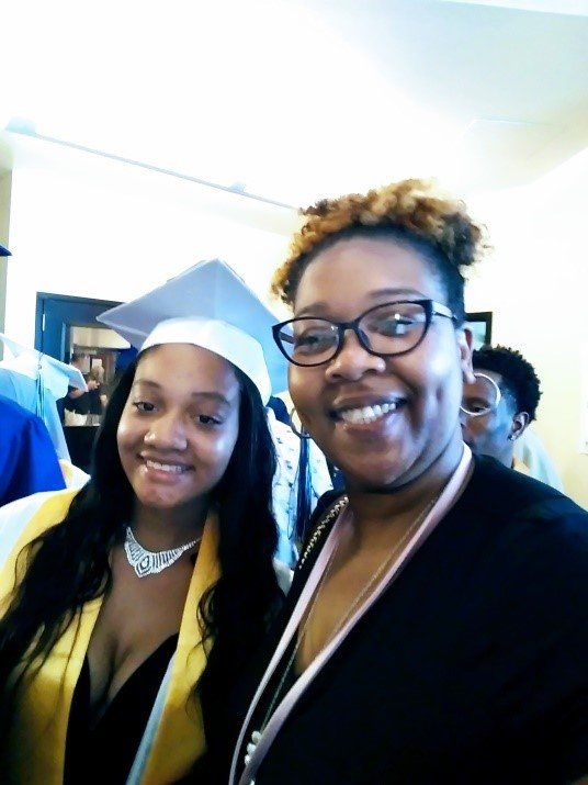 Woman with a graduation cap and gown smiles with a woman wearing glasses, both indoors.