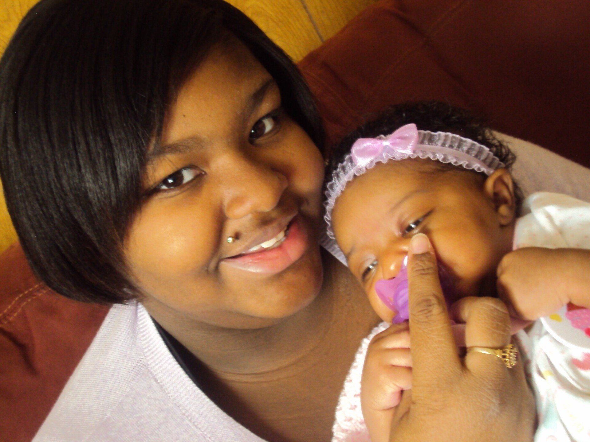 Young woman holding a baby; woman smiles, points at baby's face. Baby wears headband and has pacifier. Brown background.