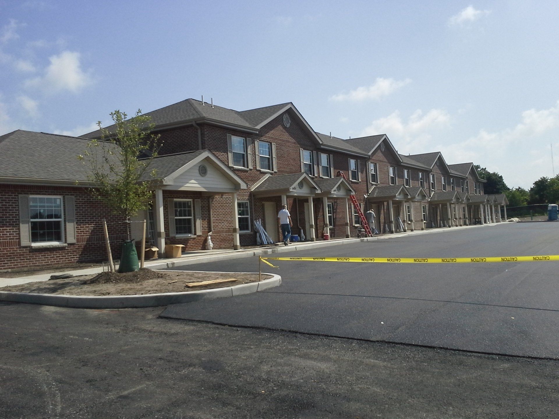 Row of brick townhouses with a newly paved parking lot, cordoned off by caution tape, on a sunny day.