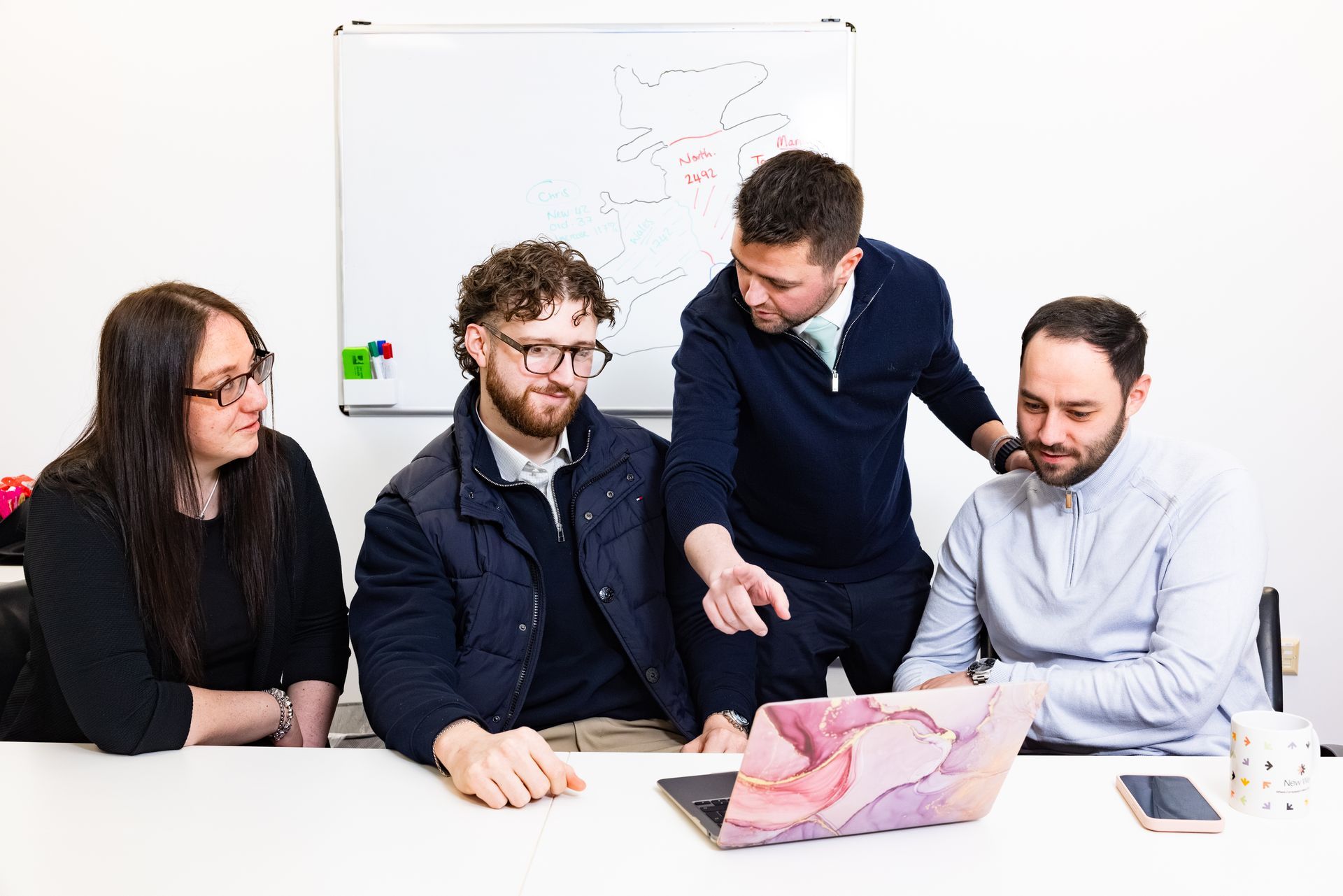 Four coworkers gathered around a laptop, reviewing a map and discussing work in a bright office.