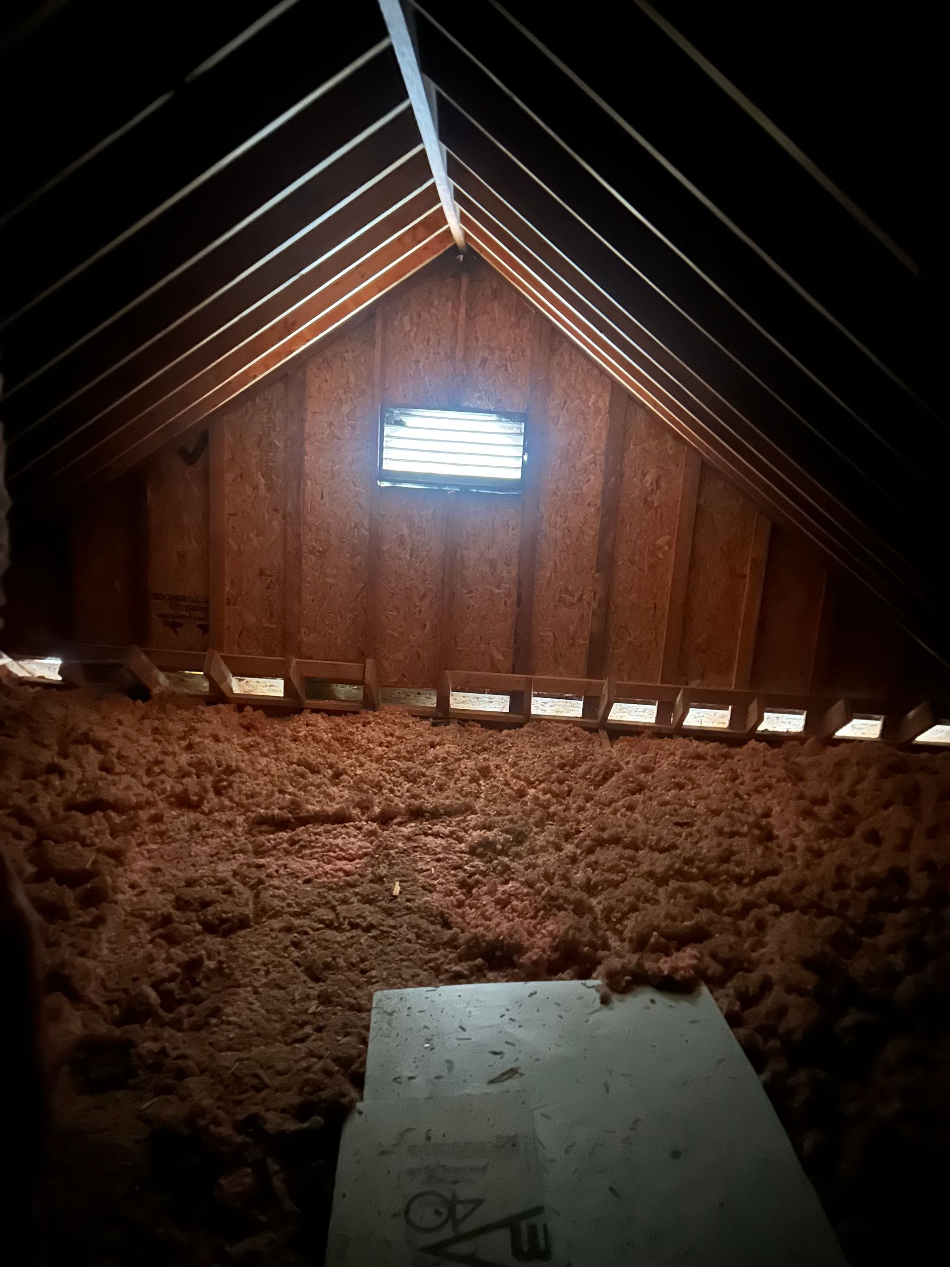 Attic with brown insulation, wood beams, a small window, and a wood plank.