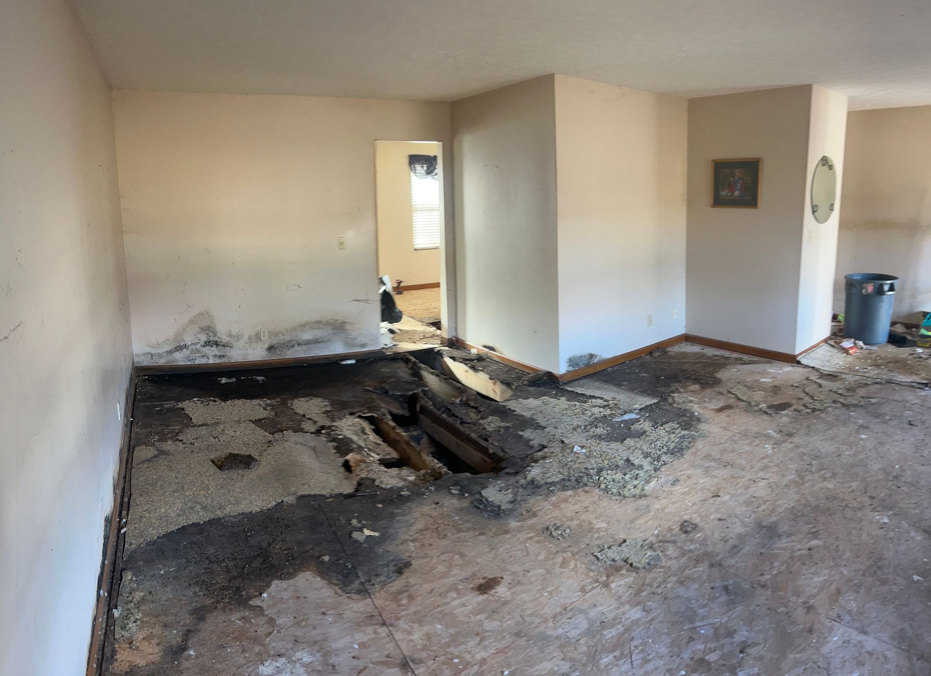 Damaged interior room: hole in floor, charred area, damaged walls, door in background.