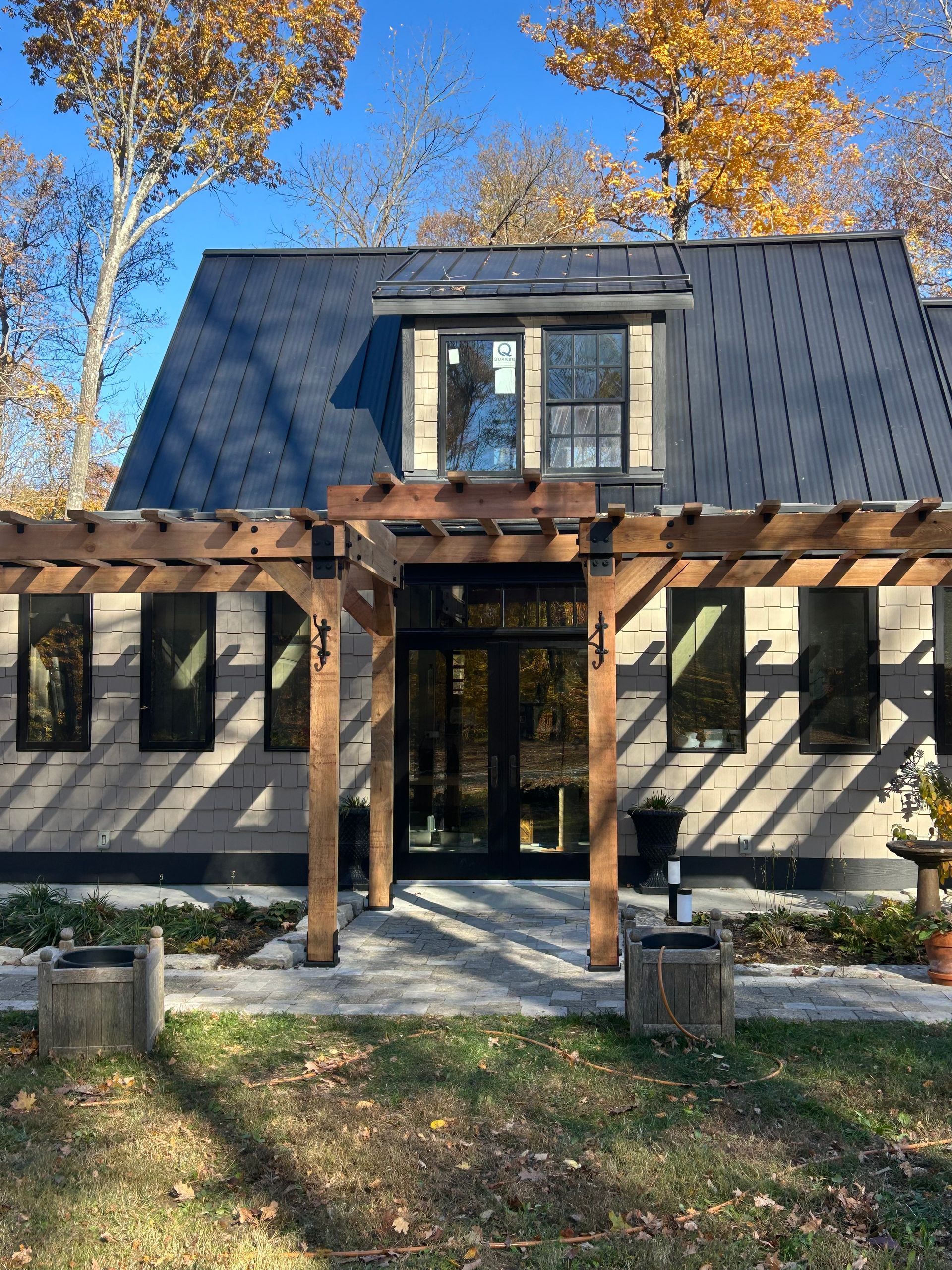 Modern house with black roof, wooden pergola, and stone walkway. Autumn trees in the background.