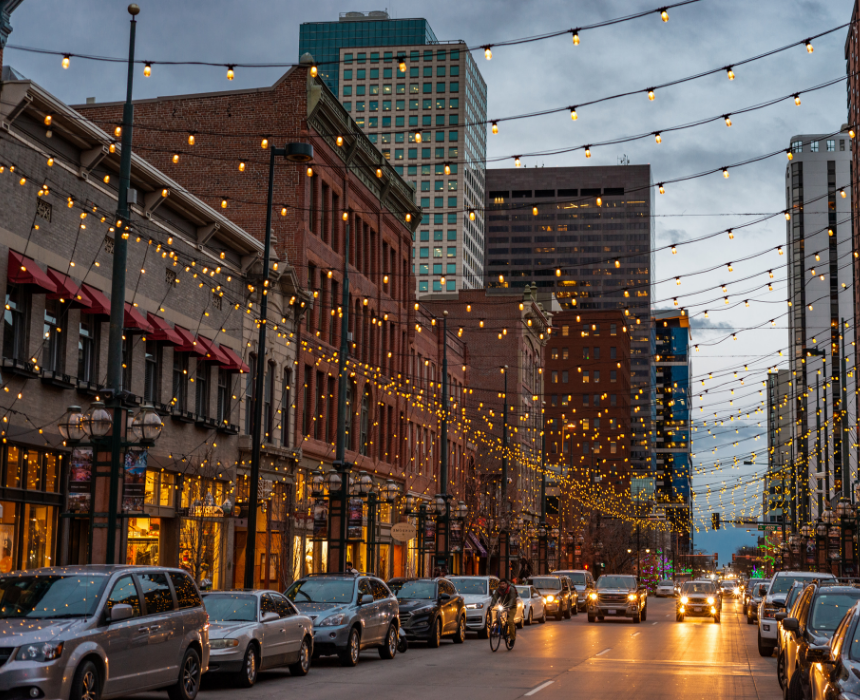 A city street with cars parked on the side of it and christmas lights hanging from the buildings.