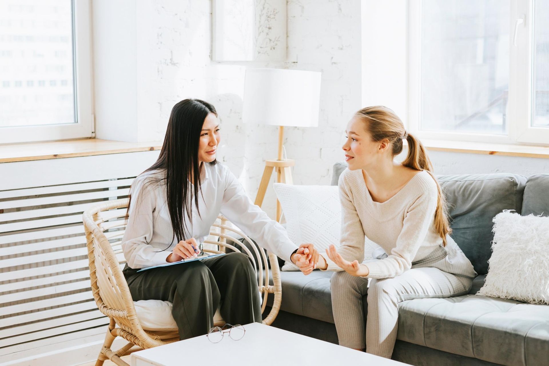 A therapist consoles a client in a light-filled room. The therapist holds the client's hand while they talk.