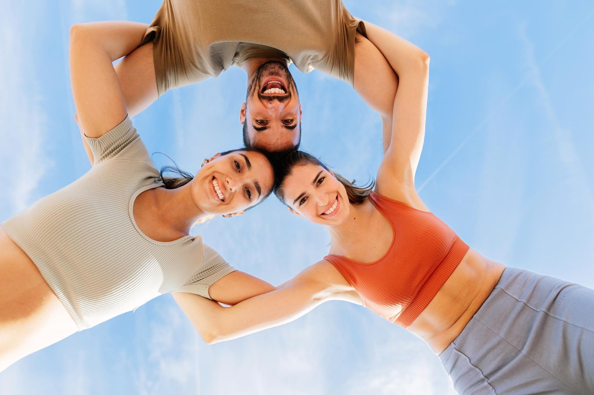 Three smiling people, heads together forming a circle, arms interlocked against a blue sky.
