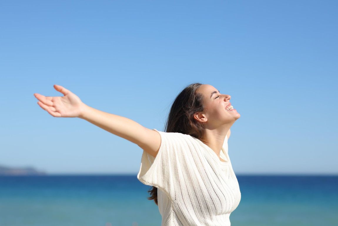 Woman with arms outstretched, smiling in front of ocean and blue sky.