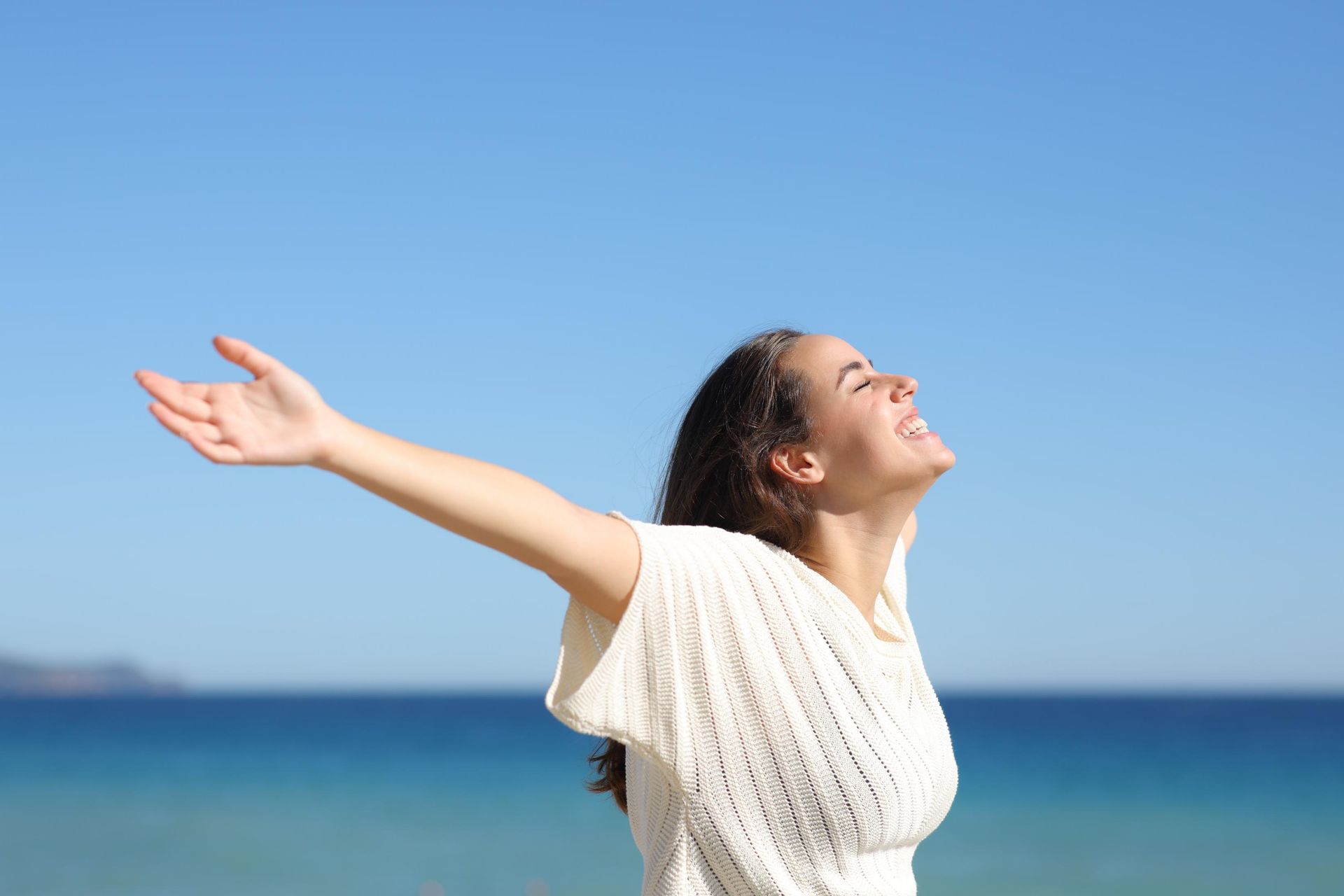 Woman with arms outstretched, smiling in front of ocean and blue sky.