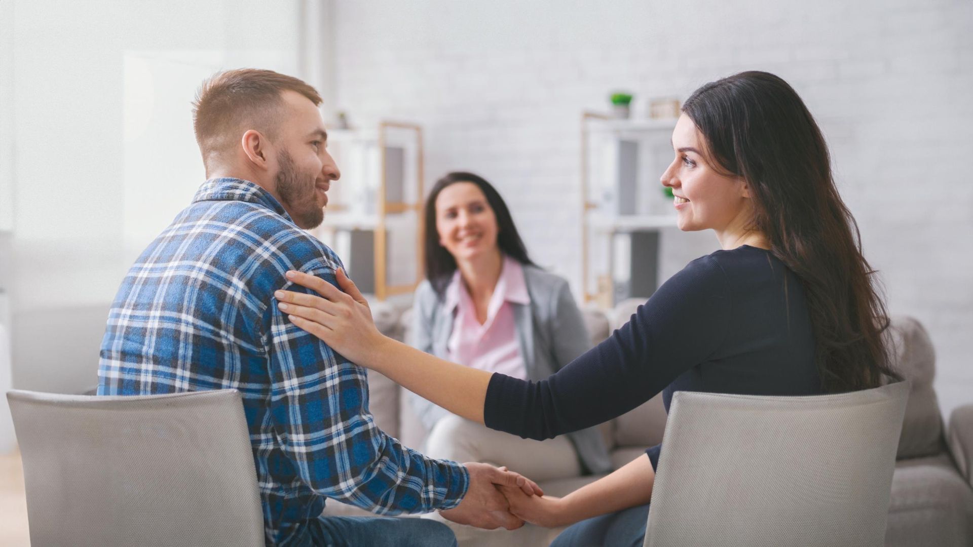 Couple in therapy, holding hands and smiling, counselor in background.