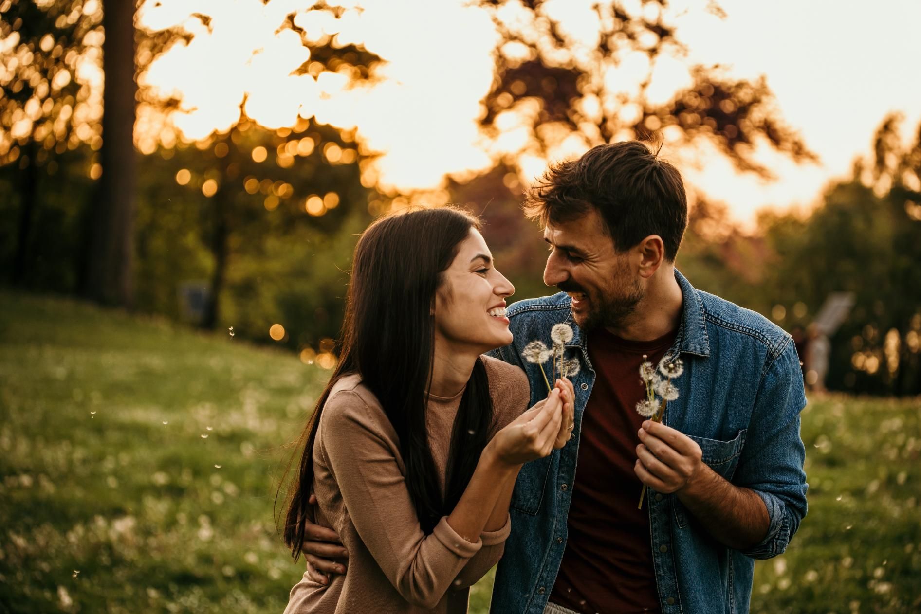 Couple smiling at each other outdoors, blowing dandelion seeds. Golden hour lighting.
