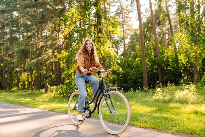 Woman riding a bicycle on a paved path in a sunny park; she wears jeans and an orange top.
