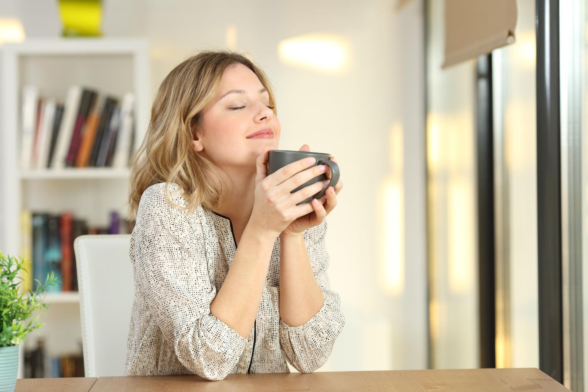 Woman smelling coffee, eyes closed, smiling. Sitting at a table near a window, books visible in background.