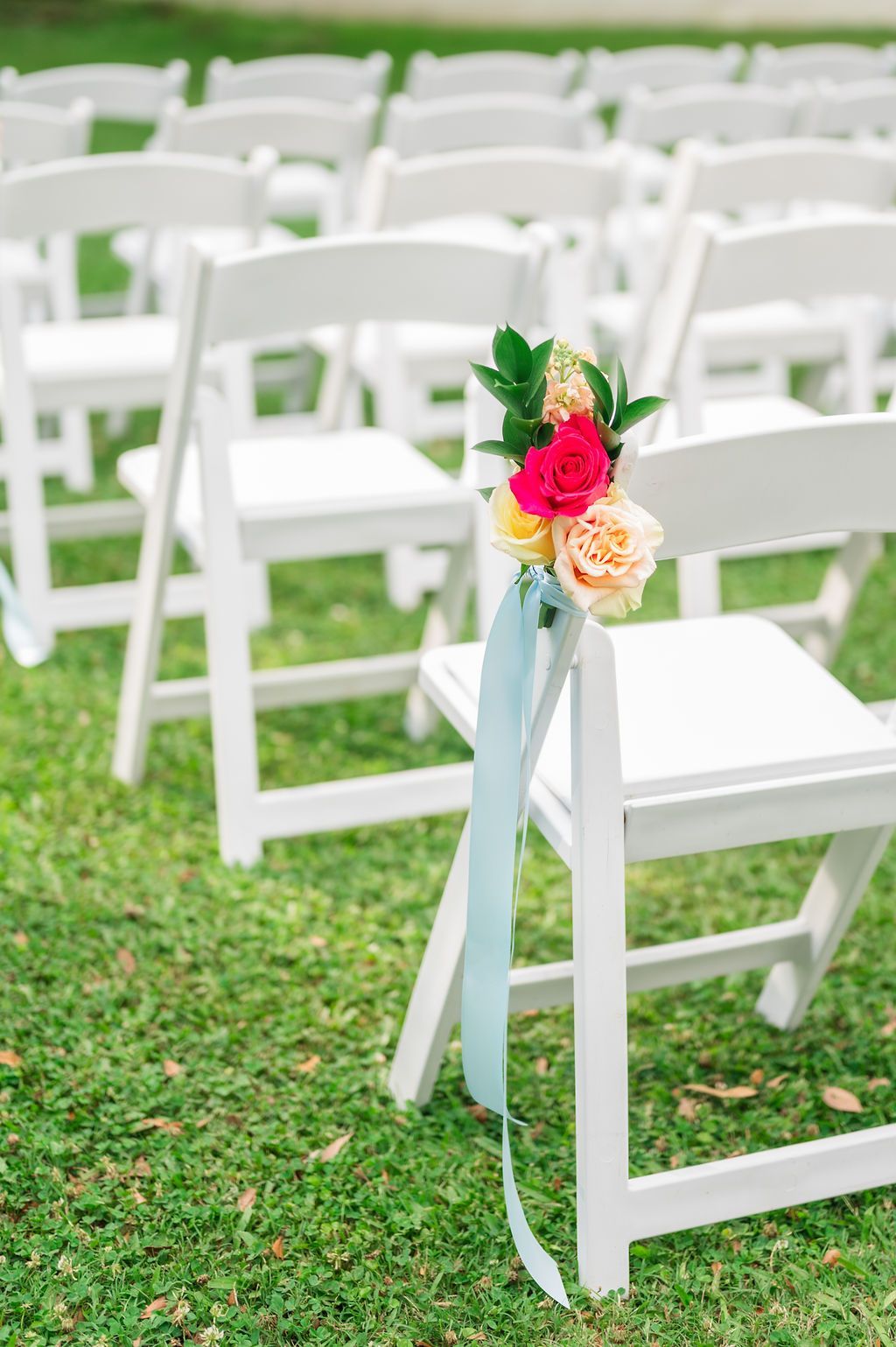 White chairs arranged on grass with floral decorations and blue ribbons.