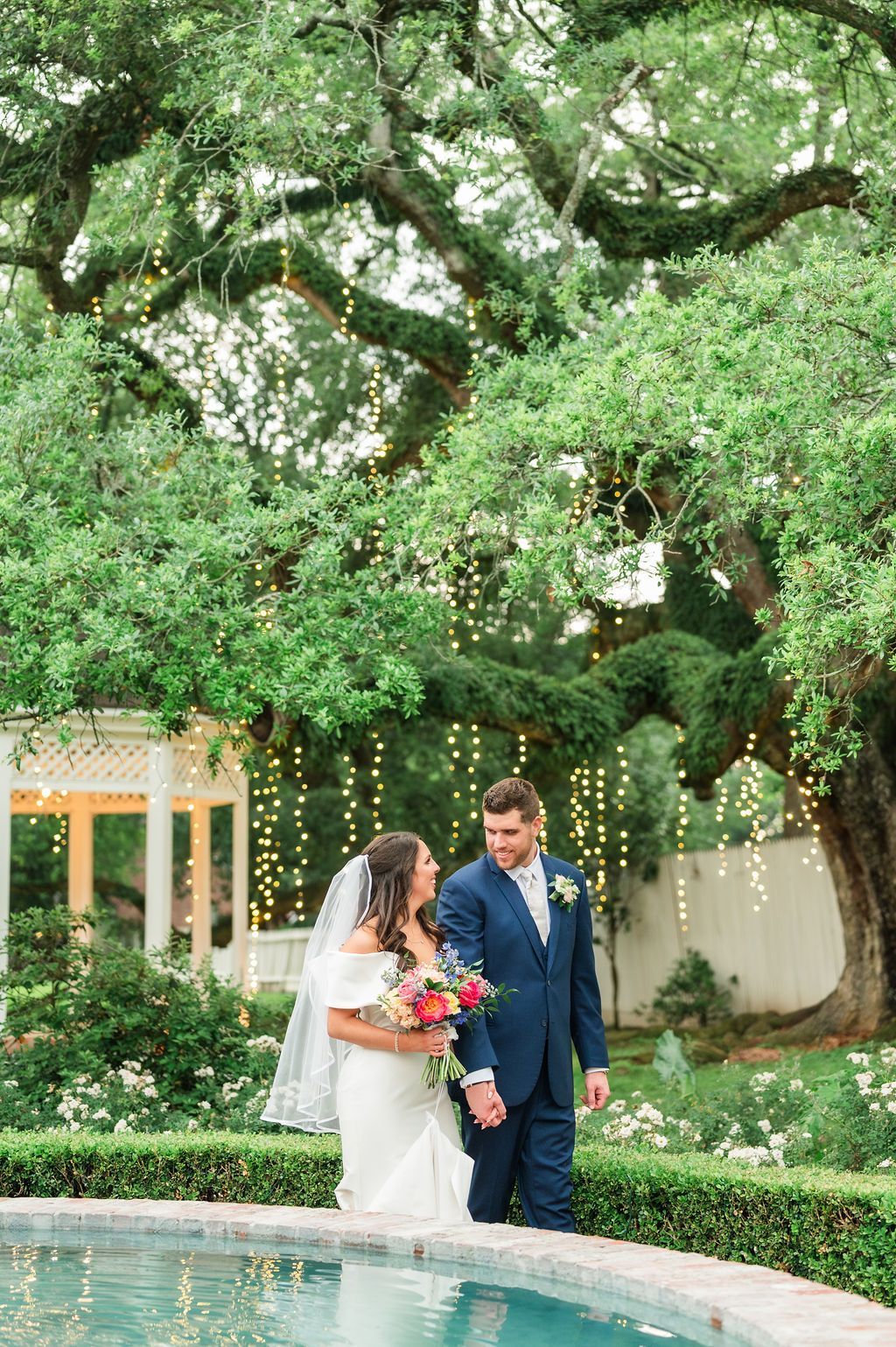 Bride and groom walk near a pool. They are under a large tree with lights.