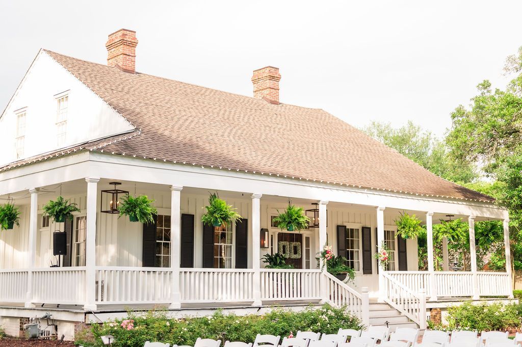White house with a long porch, brown roof, and hanging plants, likely for an outdoor event.