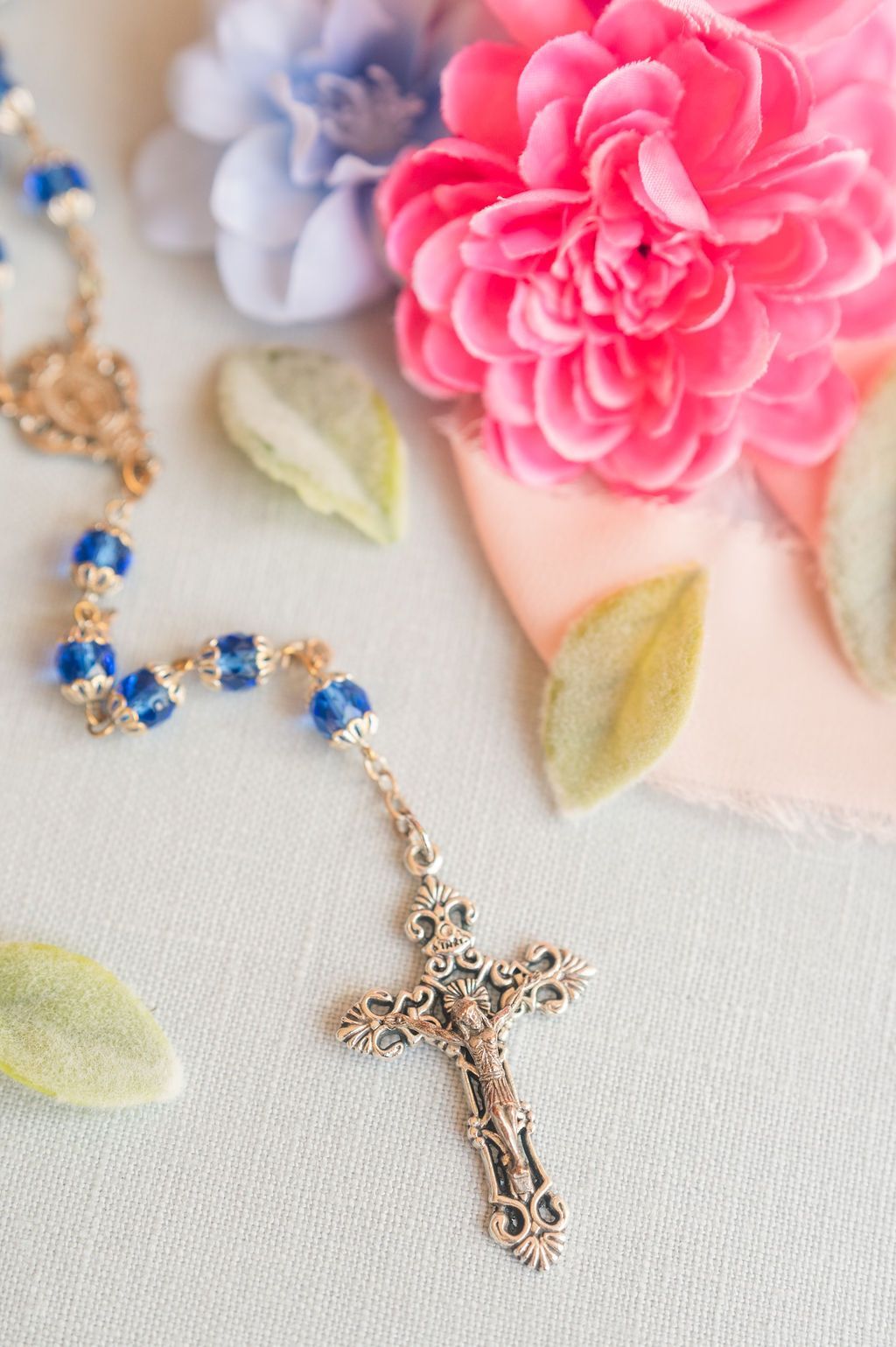 Rosary beads with blue accents and ornate cross, placed with pink and blue flowers.