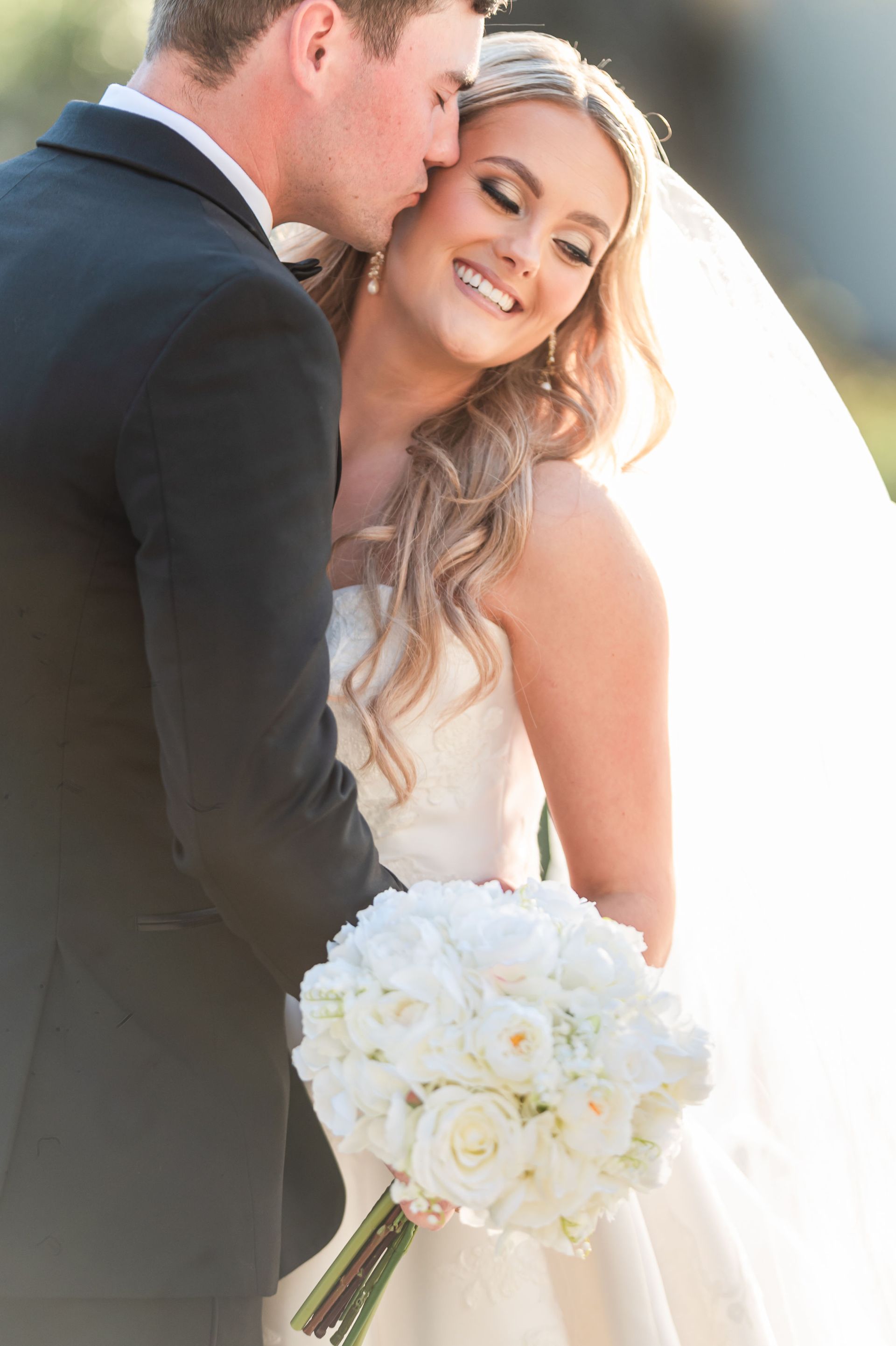 Bride in white dress smiles as groom kisses her cheek; she holds white rose bouquet.