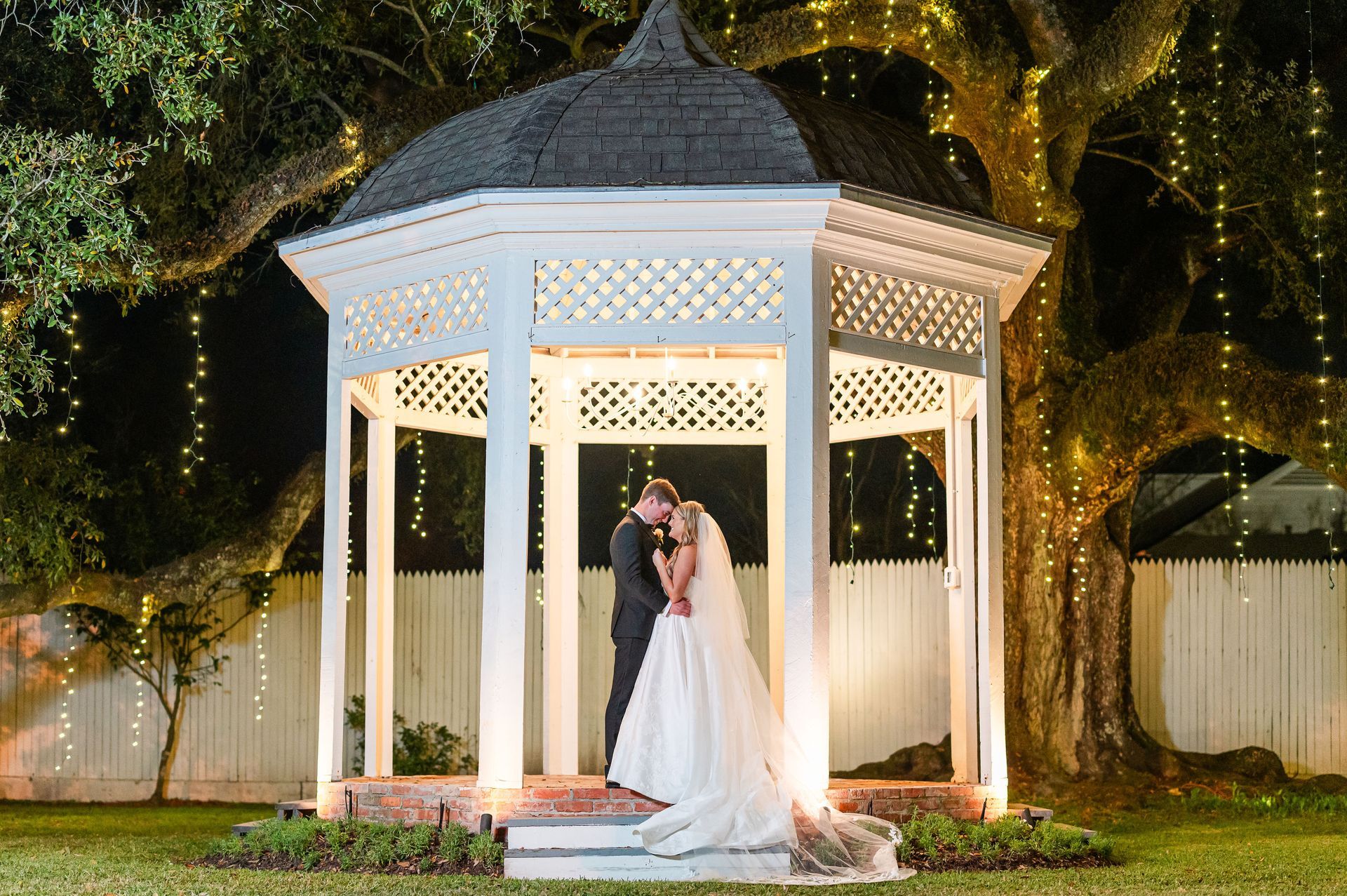 Couple kissing in a white gazebo, illuminated by string lights, beneath a large tree in a garden setting.
