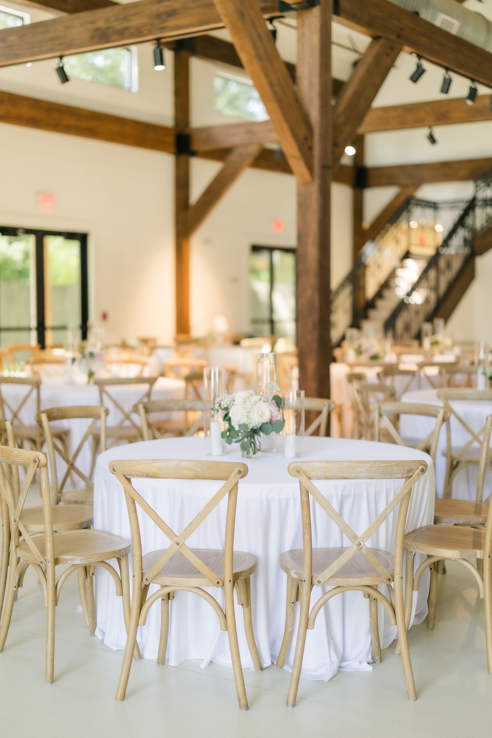 Round tables set for a celebration in a bright, wood-beamed room. White linens, cross-back chairs.
