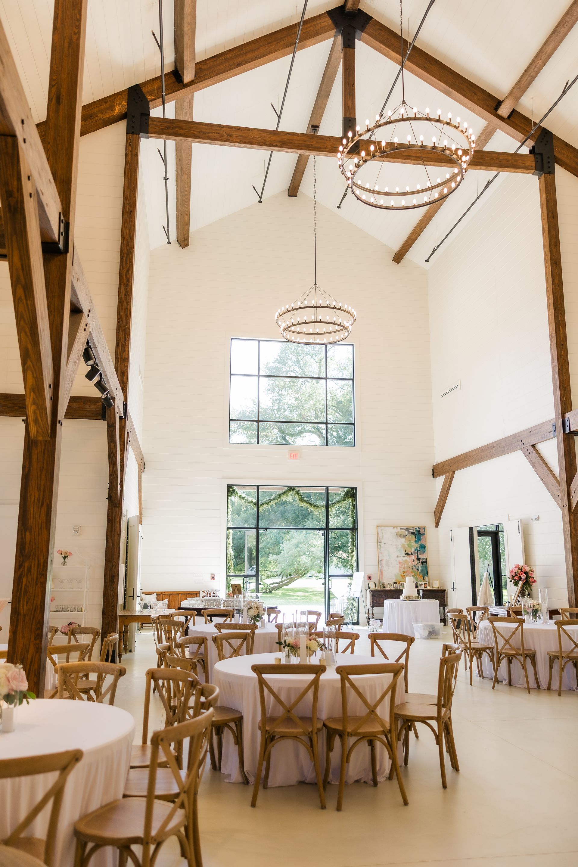 Wedding reception hall with high ceilings, wooden beams, round tables, and cross-back chairs.
