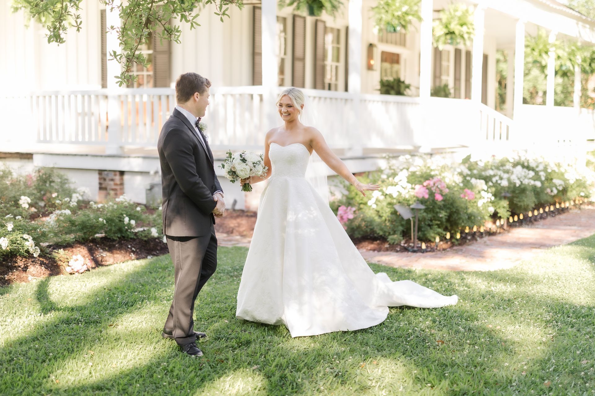 Bride in white dress smiles at groom in suit; garden, white house background.
