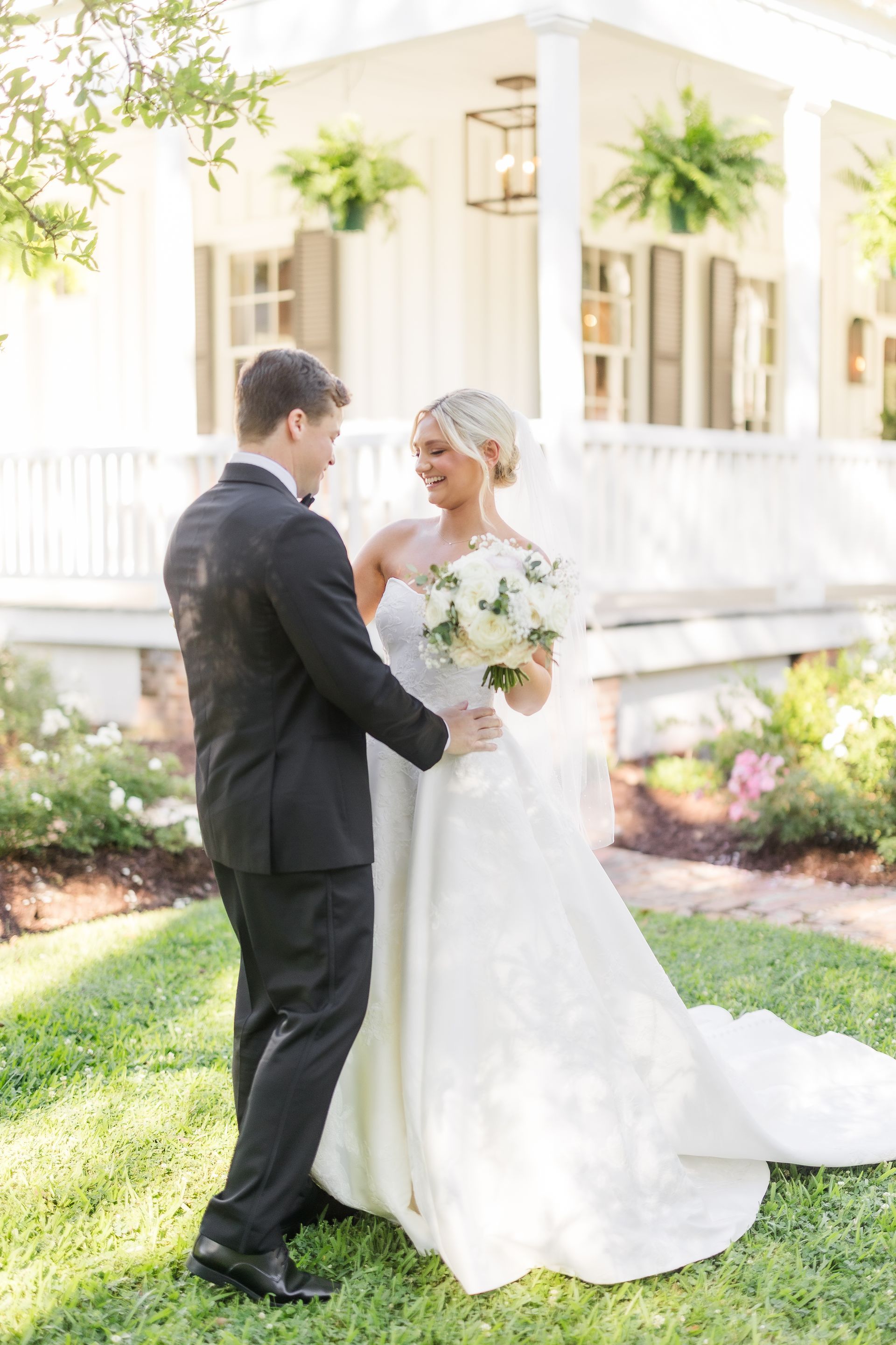 Bride and groom embrace outdoors, holding flowers, in front of a white building with plants.