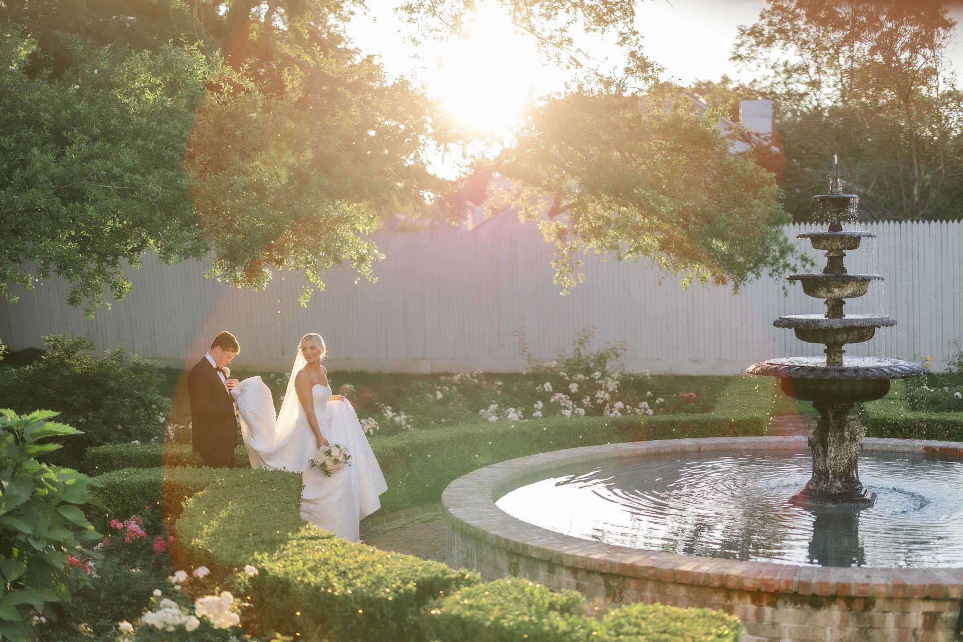 Bride and groom walk near a fountain in a garden; sunlight streams through trees.