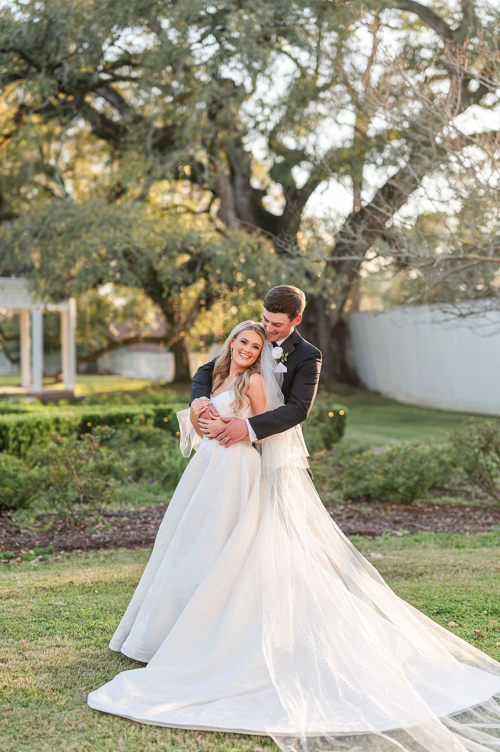 Couple embraces, bride in a white gown and veil, groom in a black suit, outdoors near a large tree.
