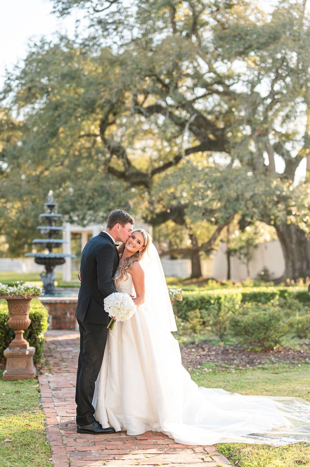 Bride and groom embrace, leaning in for a kiss in a garden. Bride wears a long veil.