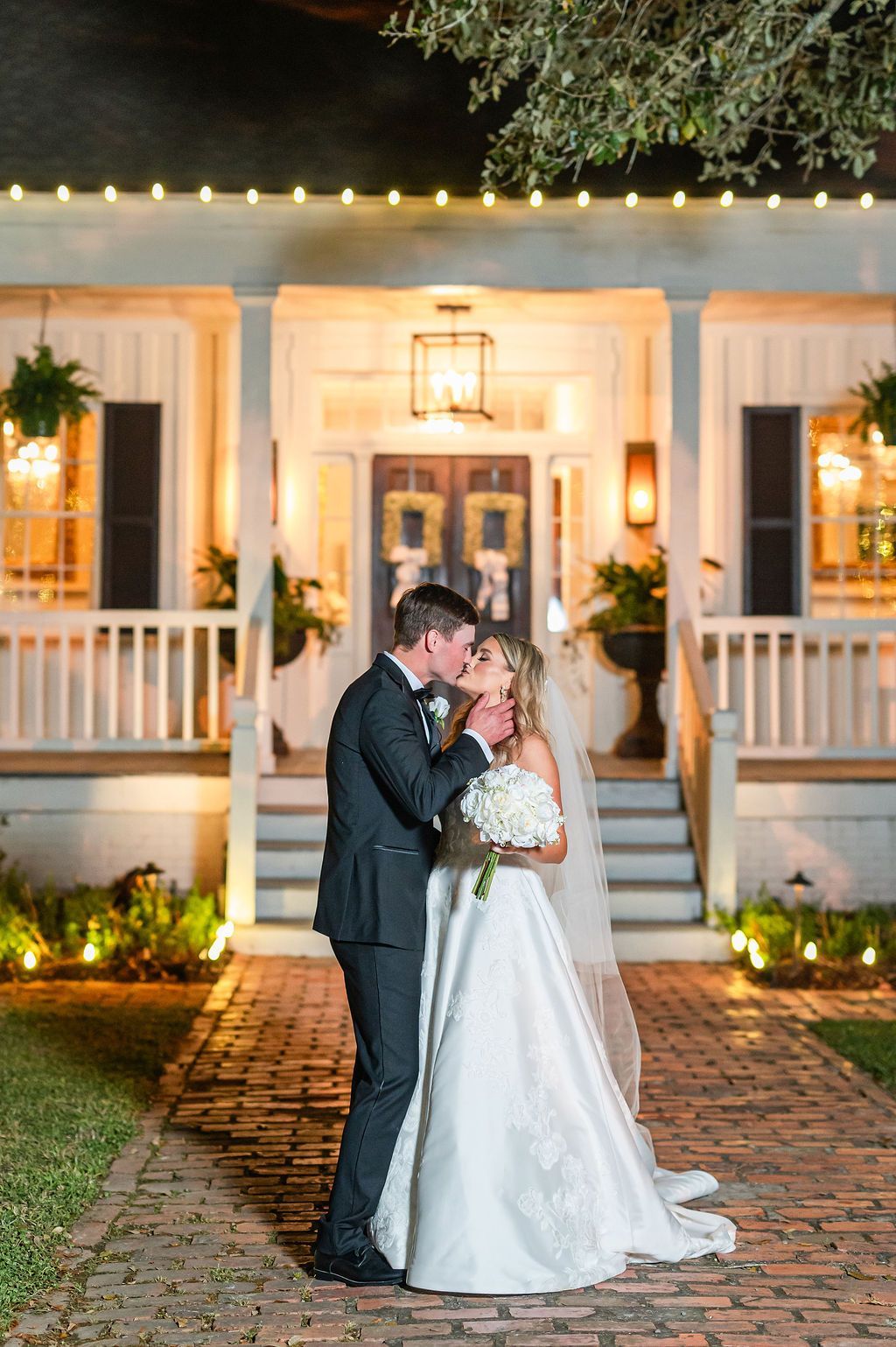 Newlyweds kissing on brick path in front of a house, lit with string lights.