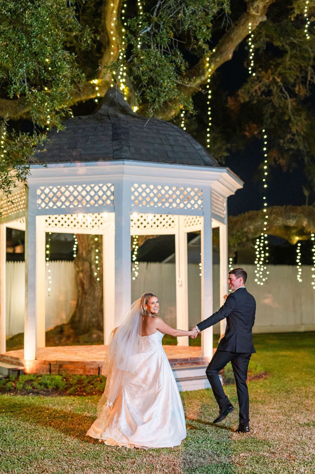 Bride and groom dance in a garden at night near a gazebo lit with string lights.