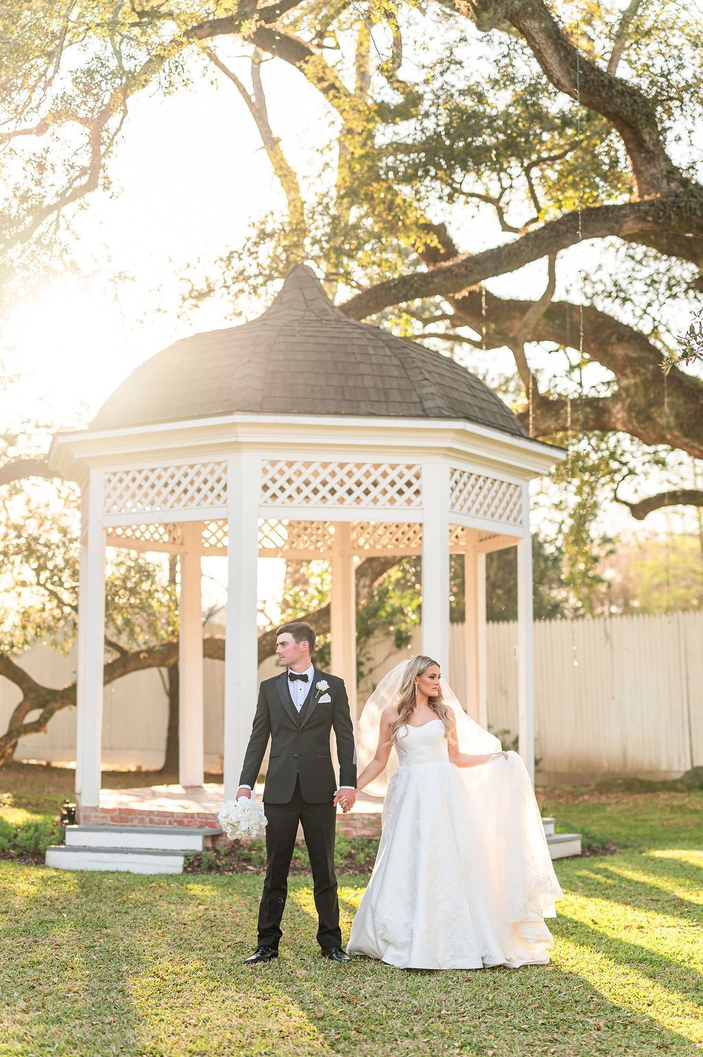 Couple holding hands in front of white gazebo. Bride in white gown, groom in suit, sun shining.