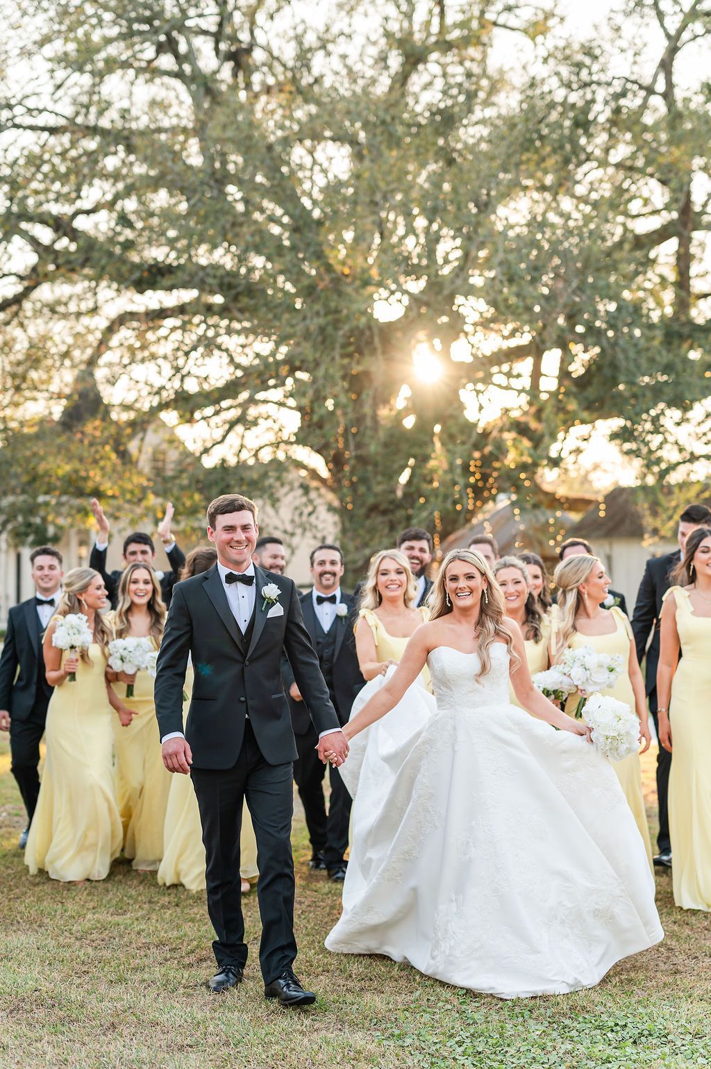 Bride and groom lead wedding party, smiling, outdoors; bridesmaids in yellow dresses, groomsmen in black tuxedos.