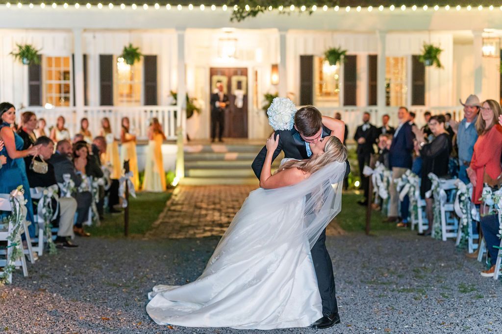 Bride and groom share a celebratory dip outdoors. Guests watch from rows of chairs near a white building with lights.