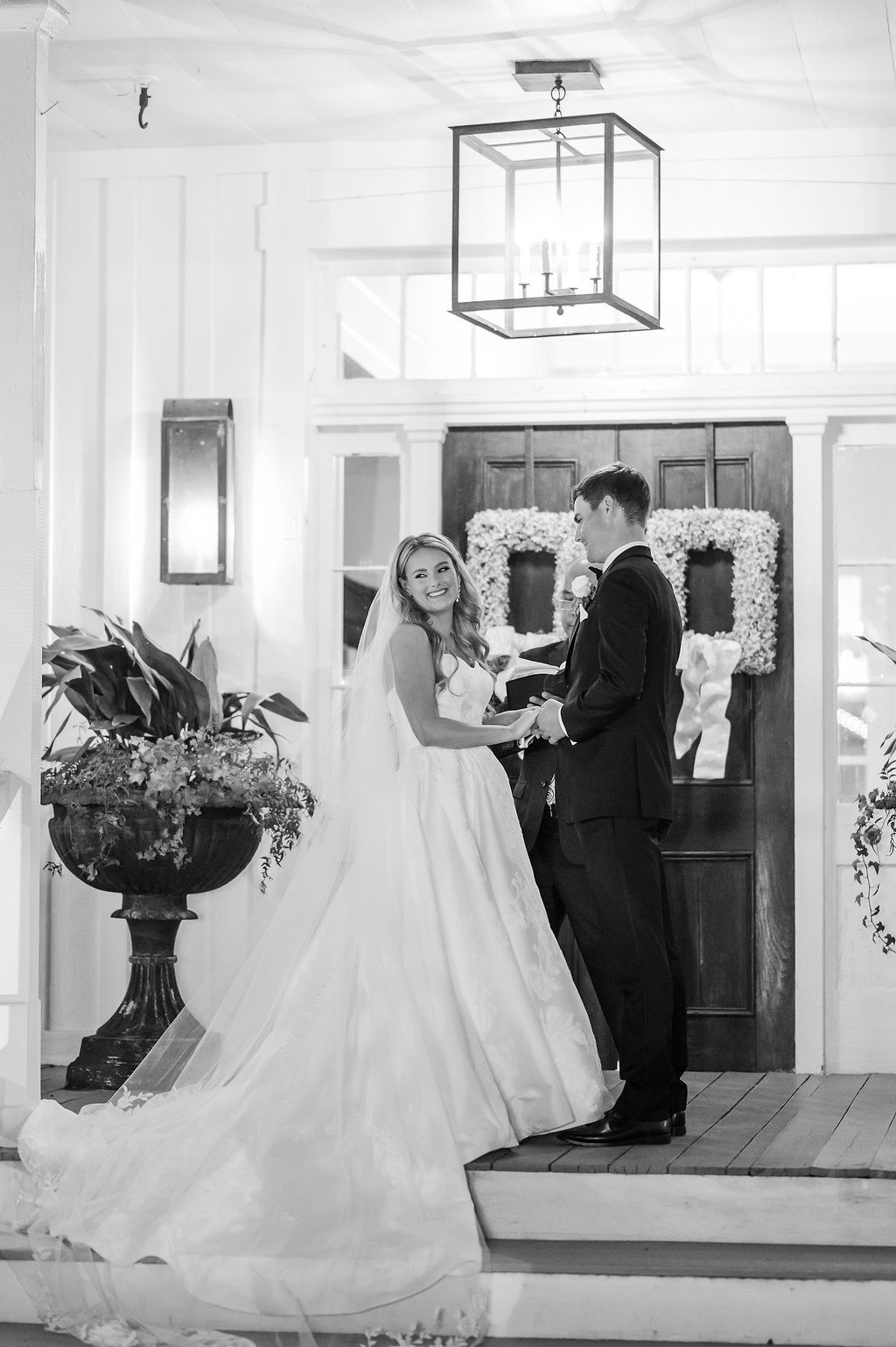 Bride and groom holding hands, standing on porch, smiling. Wedding dress, dark suit, wreath on door.