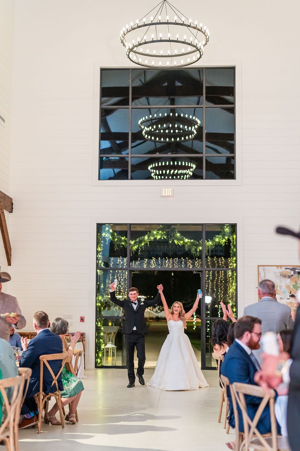 Newlyweds enter a wedding reception, arms raised, celebrating. Bright room, guests seated at tables.