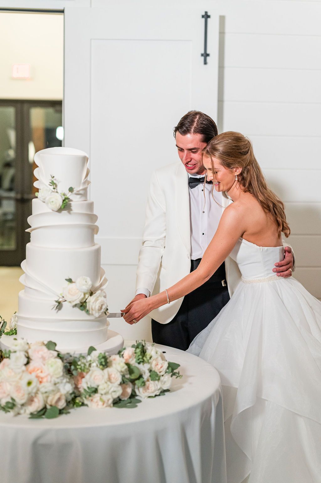 Bride and groom cutting wedding cake. White tiered cake with floral accents. Couple smiles in front of white wall.