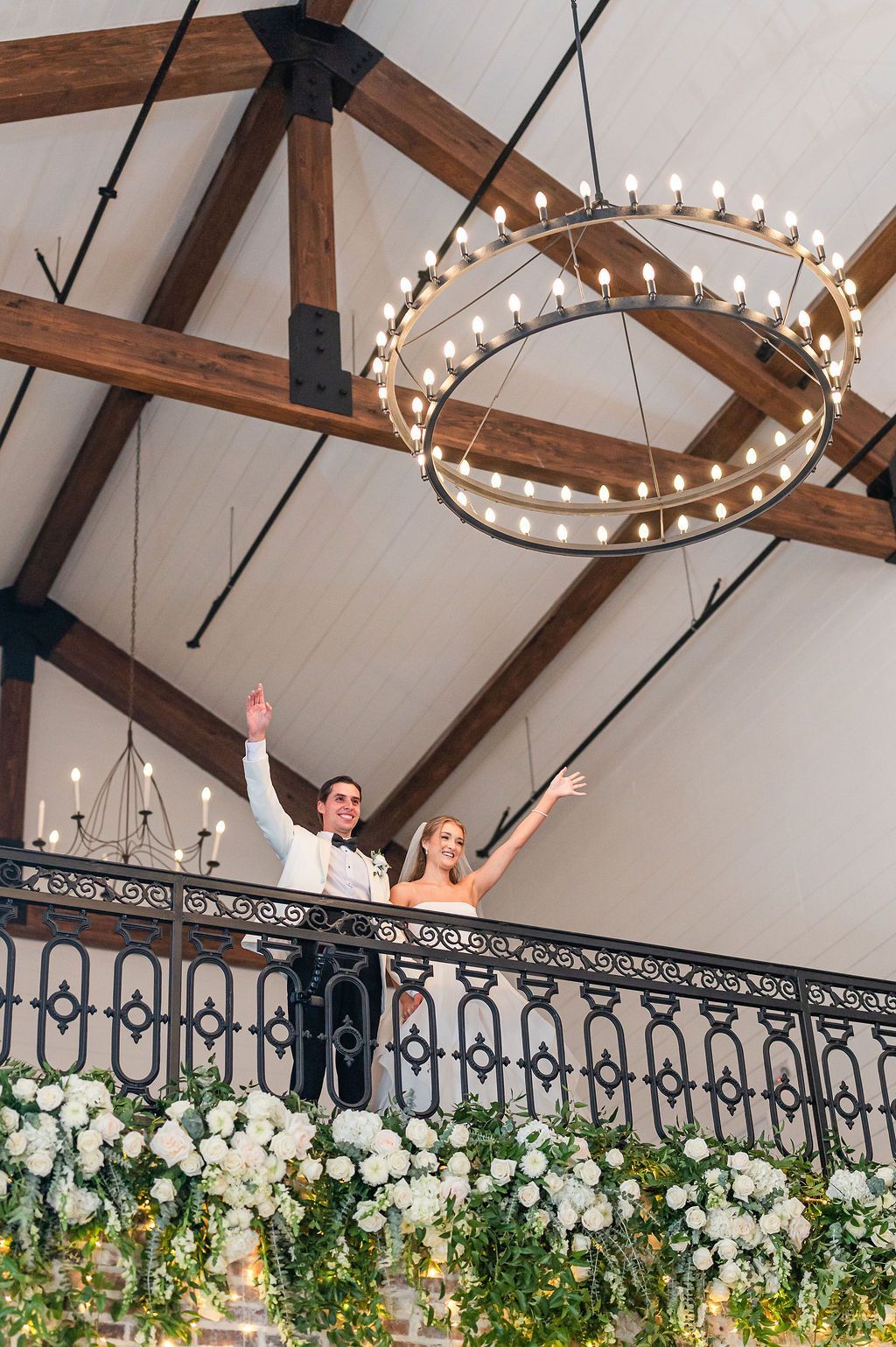 Newlyweds wave from a balcony decorated with white flowers. Bright chandelier above.
