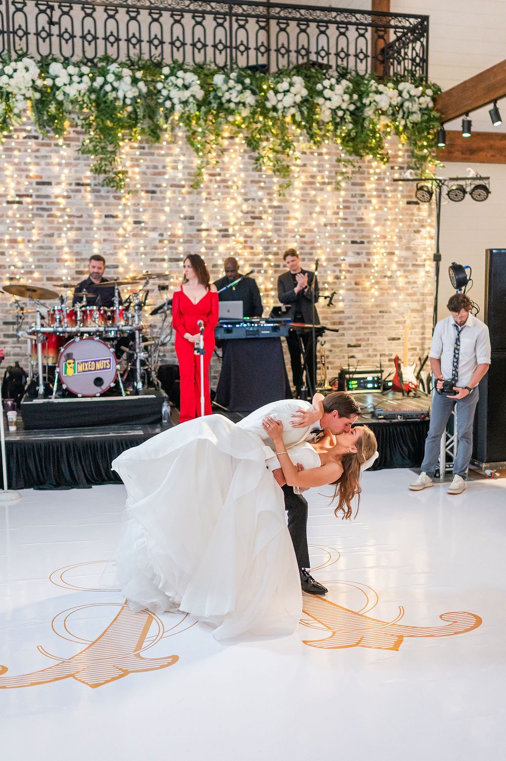 Bride and groom dance, band performs at a reception with floral and brick background, and twinkle lights.