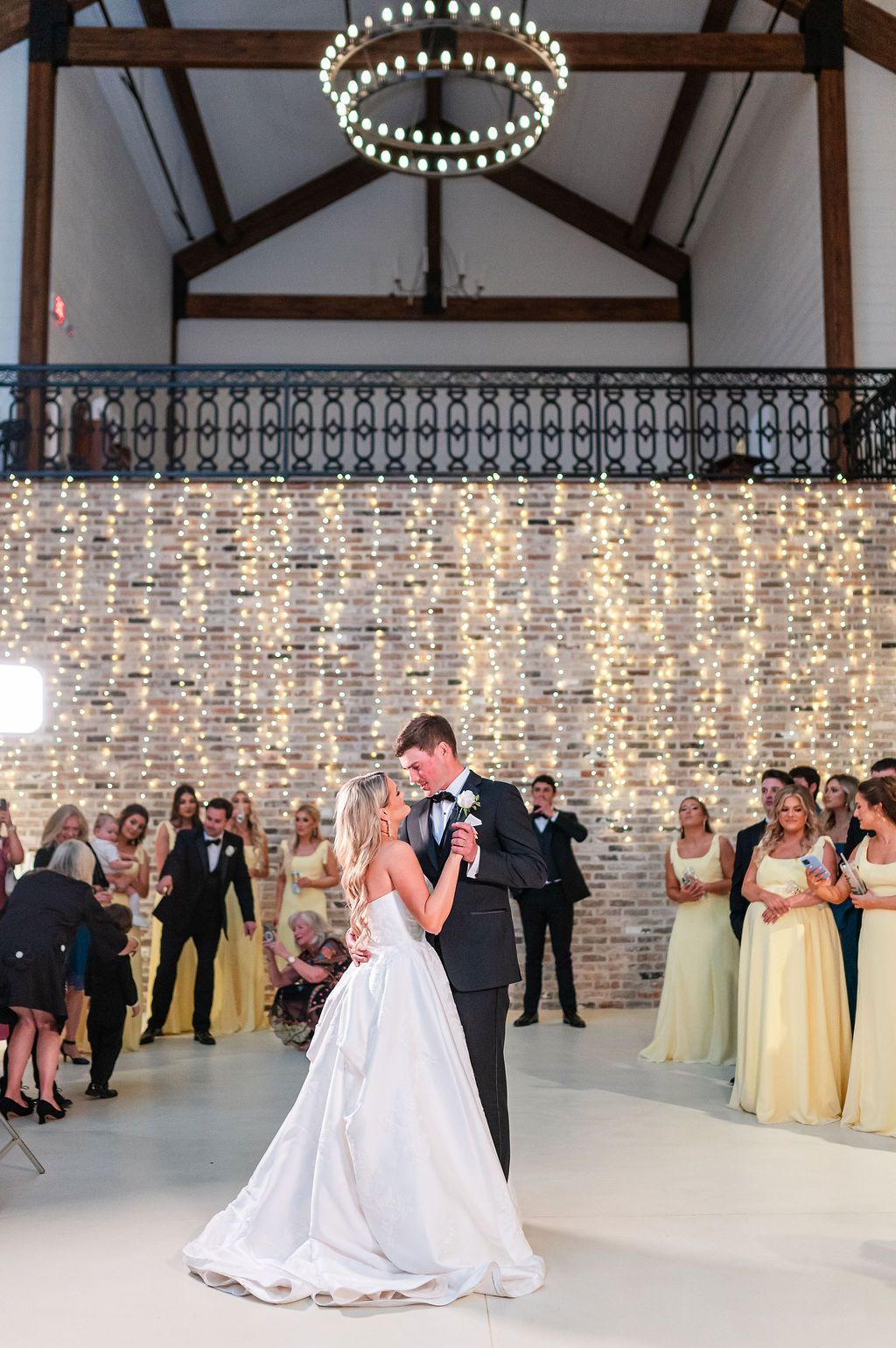 Bride and groom dance at reception; guests watch. Brick wall backdrop with string lights, chandelier overhead.