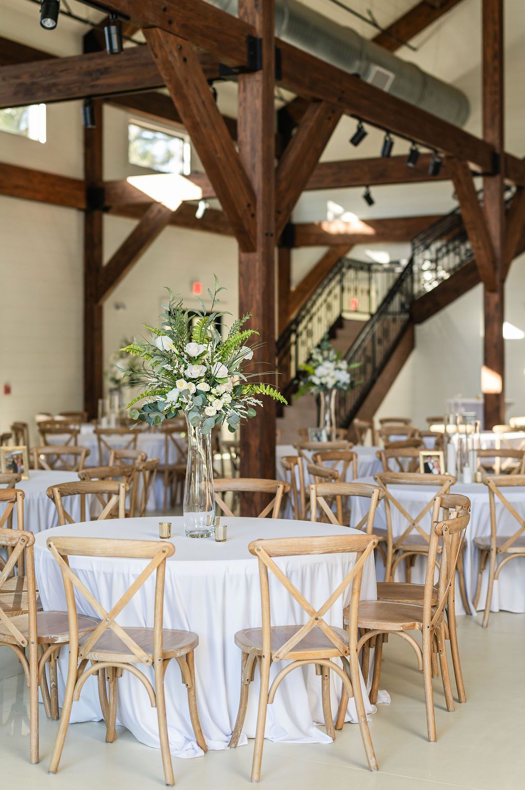 Indoor reception: Round tables with white linens, wooden chairs, floral centerpieces, exposed beams, and a staircase.