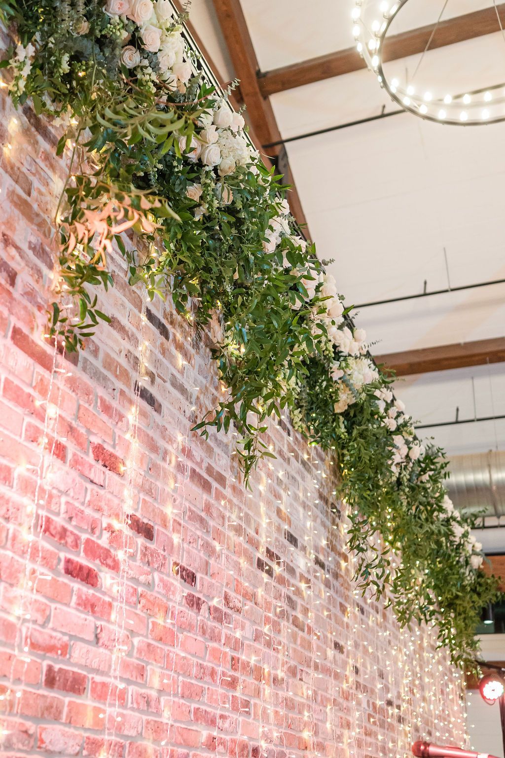Brick wall decorated with white flowers, green vines, and string lights, indoors.