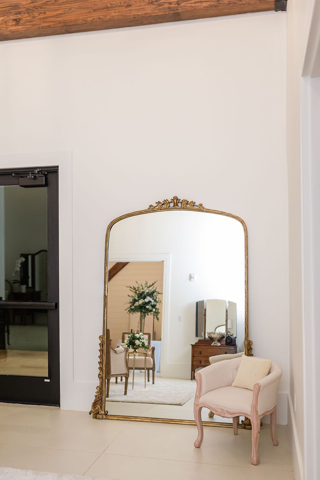 Large ornate gold mirror reflects room with chair and floral arrangement. White walls and wooden beam.