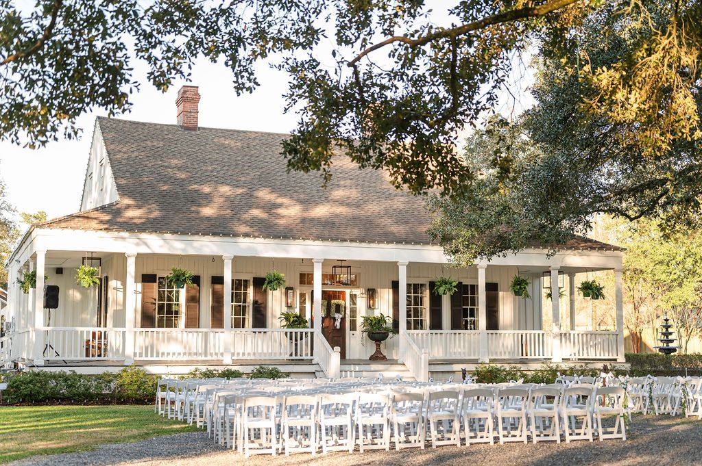 White house with porch, outdoor wedding setup: chairs face the building, green grass, trees.